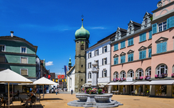 Stadtplatz mit historischen Gebäuden und einem zentralen Brunnen bei klarem Himmel.