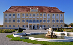 Barockschloss mit symmetrischem Garten und Brunnen im Vordergrund.