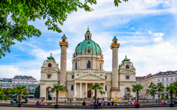 Barockkirche mit grüner Kuppel und zwei Säulen, umgeben von Bäumen und Leuten.