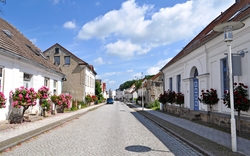 Gepflasterte Straße mit alten Häusern und blühenden Blumen unter blauem Himmel.
