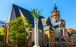 Kirche mit markanter Säule im Vordergrund vor blauem Himmel.